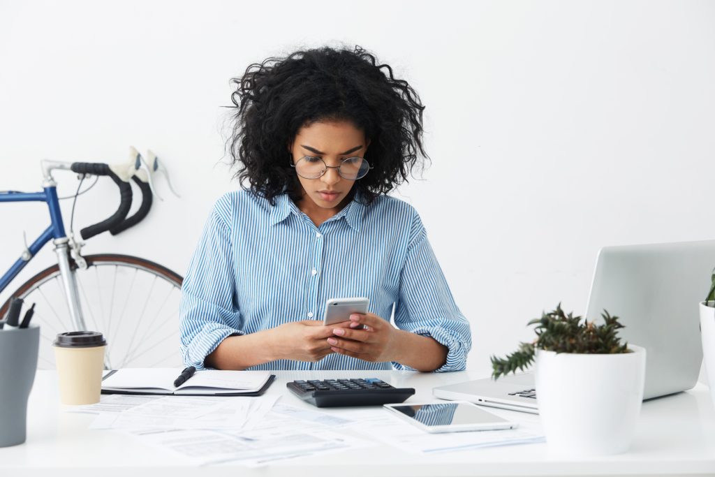 People and modern technologies concept. Concentrated young mixed race female secretary reading urgent message on cell phone while sitting at her workplace with electronic gadgets and papers